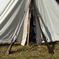 A canvas tent is set up on a grassy area, with its entrance open. Two rifles, reminiscent of reenactments like Dunamunde 2010, are propped up against the tent, crossing each other at the entrance. The ground around the tent is grassy, showcasing a rustic outdoor setting. The interior of the tent is not visible.