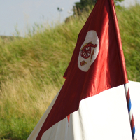 A red flag, adorned with a white badge featuring a medieval knight's helmet and crossed anchors, is mounted on a striped pole. This scene evokes the 2010 Dunamunde festival, set against a backdrop of green vegetation and a clear sky in front of a white tent pitched in a grassy area.