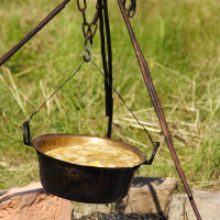 A cast-iron pot filled with food hangs over a fire pit, reminiscent of Dunamunde 2010. The pot is suspended by chains attached to a metal tripod, while ashes and small burned wood pieces scatter below. The grassy terrain in the background sets the scene for outdoor cooking or camping.