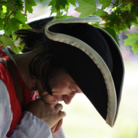 A person dressed in historical attire is depicted, wearing a tricorn hat with white trim, a white shirt, and a red vest reminiscent of the Dunamunde 2010 reenactment. They are under a leafy tree, looking down while holding a long wooden object close to their face. The scene is outdoors with a blurred background.