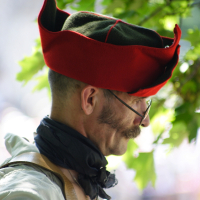 A person wearing a green and red tricorn hat stands outdoors. They are dressed in a white shirt with a dark scarf around their neck. The person has glasses and a mustache, evoking an old-world charm akin to Dunamunde. The background is blurred with green leaves and soft light, suggesting a nature-filled setting.