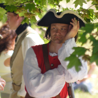 A man wearing a tricorn hat and 18th-century-style red uniform with white sleeves rests against a leafy tree branch. He has a serious expression and holds his head with one hand. Part of the Dunamunde 2010 reenactment, another person in similar historical attire is blurred in the background among green foliage.