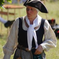 A person dressed in historical attire stands outdoors. They wear a black tricorne hat, a white headscarf, a dark vest over a beige long-sleeve blouse, and a striped apron. Holding a green pouch, they appear to be part of the Dunamunde 2010 reenactment, with blurred people and tents in the background.