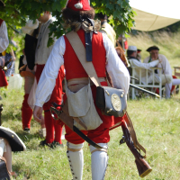 A person dressed in historical military attire, reminiscent of Dunamunde 2010, including a red coat, white pants, tall boots, and a red hat, walks away from the camera on a grassy field. The person carries a musket and a satchel while other similarly dressed individuals are seated in the background under a tree.