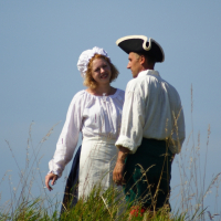 Two people dressed in historical costumes are standing in a grassy field under a clear blue sky. The woman, smiling, is wearing a white bonnet, blouse, and apron, while the man is in a white shirt and tricorn hat. They appear to be reenacting an event from Dunamunde in 2010 as they engage in conversation.