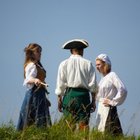 Three people dressed in historical clothing stand on a grassy hill against a clear blue sky. One woman in a blue skirt and brown vest holds a scroll, while another in an apron and bonnet interacts with them. A man in a tricorn hat and green pants stands between the two women. Tall grass and wildflowers surround them, evoking the spirit of Dunamunde despite the year being 2010