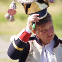 A man dressed in a historical Dunamunde military uniform, featuring a dark jacket with red and white cuffs, gold buttons, and a cream-colored vest. He is lifting a tall, ornate hat with gold embellishments, tassels, and the number "1" on it. The background is blurred, showing outdoor greenery.