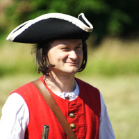 A man in historical attire stands outdoors on a sunny day, wearing a black tricorn hat with white trim, a red waistcoat with brass buttons over a white long-sleeved shirt, and holding a musket. The background is blurred with greenery. He is smiling and looking into the distance, reminiscent of Dunamunde reenactments from 2010.