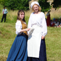 Two women stand outdoors in traditional historical attire. The woman on the left wears a blue skirt, white blouse, and brown bodice, holding a white fan. The woman on the right, dressed in a white blouse with puffed sleeves, long dark blue skirt, white apron, and bonnet, evokes the spirit of Dunamunde amidst the 2010 summer greenery.