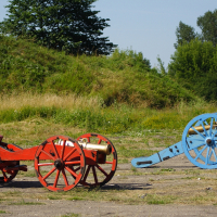 Two colorful cannons are situated outdoors on a sunny day. The cannon on the left is painted red with yellow accents, while the one on the right is blue with golden details. They are displayed on a paved surface with lush, grassy mounds and trees in the background, reminiscent of Dunamunde 2010.