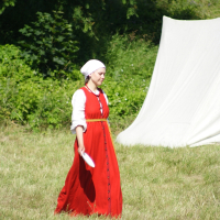 A woman in historical costume walks on grass at what appears to be a 2010 reenactment event. She wears a bright red dress with long sleeves and a white headscarf, holding a folded white paper in her right hand. Behind her, greenery and a white cloth setup, possibly a tent, completes the scene reminiscent of Dunamunde.