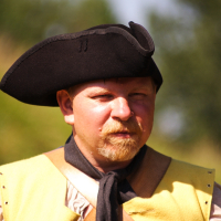 A man dressed as a historical reenactor for Dunamunde 2010 wears a tricorne hat, a yellow vest over a white shirt, and a brown leather strap across his chest. He has a light beard and stands against an outdoor background blurred with greenery. The expression on his face is serious and focused.