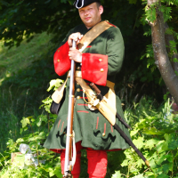 A man dressed in historical military attire stands in a lush, green setting, reminiscent of Dunamunde 2010. He wears a dark green coat with red cuffs and trim, red pants, and a tricorn hat. Holding a long rifle with a beige strap across his chest, the background features green foliage and a partially visible tree trunk.