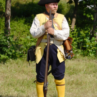 A man dressed in colonial-era clothing stands in a grassy field at the Dunamunde reenactment. He wears a tricorn hat, a light-colored vest over a white shirt, dark blue trousers, and yellow socks. He holds a long rifle upright in front of him and has various pouches and equipment strapped to his waist. Trees form the backdrop.