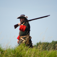 A man dressed in historical military attire, reminiscent of Dunamunde 2010, consisting of a black hat, dark green coat with red sleeves, and beige sash, walks outdoors. He carries a long rifle over his shoulder. In the background, there is tall grass and a clear blue sky.