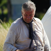 An older man with gray hair stands outdoors, wearing a light-colored long-sleeve shirt and gray overalls. He holds a wooden bowl in his right hand and has a dark scarf around his neck. The background is out of focus, showing some greenery and the vague outline of a building, reminiscent of Dunamunde 2010.