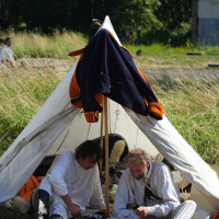 Two men dressed in white historical clothing sit under a white canvas tent pitched on grassy ground. The tent, draped with a dark cloak over the entrance, is set against a backdrop of tall grass, trees, and part of a building. Engaged in an activity, possibly writing or reading, they evoke the spirit of Dunamunde reenactments in 2010.