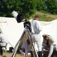 A reenactment camp scene from Dunamunde, 2010, shows four men in white historical uniforms relaxing in front of a beige canvas tent. In the foreground, several antique rifles are stacked in a teepee formation, with a black fluffy hat resting atop the rifles. Green trees and blue sky are visible in the background.