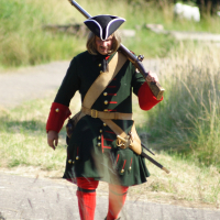 A person in a historical military uniform, reminiscent of the Battle of Dunamunde, walks outdoors on a sunny day. They wear a dark green coat with red and gold accents, a tricorn hat, red stockings, and black shoes. They carry a musket over their shoulder and a beige strap crosses their chest. Tall grass grows to either side of the path.