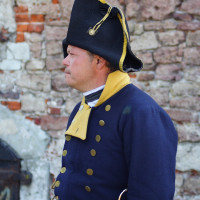 A man dressed in traditional 18th-century military attire stands against a weathered stone brick wall, reminiscent of the reenactments seen at Dunamunde 2010. He wears a navy blue coat with brass buttons, a yellow collar, and a distinctive black tricorn hat. A white cloth sash is tied around his waist, and a sheathed sword hangs by his side.