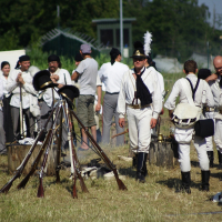 A group of reenactors dressed in historical military uniforms, including white shirts, black hats, and boots, stand in a campsite with canvas tents. Several rifles are stacked in a tripod formation in the foreground as part of the Dunamunde 2010 event, and people are engaged in various activities around the site.