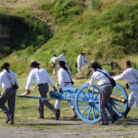 A group of men dressed in white shirts, gray pants, and black caps are pushing a light blue cannon with yellow wheels across a grassy field at Dunamunde 2010. In the background, several other people in similar attire are scattered around, with some standing near a grassy embankment and a small brick structure.