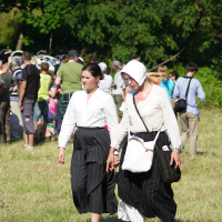 Two women dressed in historical attire walk hand-in-hand through a grassy area. Both wear black skirts and white tops; one has a headscarf and glasses, the other has her hair tied back. In the background, a group of people gathers, some dressed in similar outfits, suggesting the Dunamunde 2010 reenactment event.