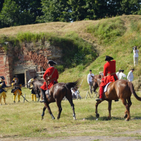 In an outdoor setting with lush greenery, two individuals in red military uniforms and black tricorn hats on horseback are riding away from a group. The group includes people in white and yellow period costumes, five parked horses, and a semi-ruined brick-and-stone structure in the background reminiscent of Dunamunde 2010.