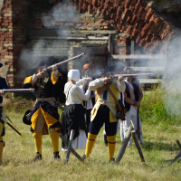 A group of historical reenactors, dressed in 18th-century military uniforms, are engaged in a mock battle at Dunamunde 2010. Four soldiers in yellow and blue uniforms are firing muskets amidst smoke. Behind them, a red brick building is partially visible, with grass and a wooden fence completing the scene.