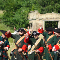 A group of costumed reenactors, dressed in historical military uniforms featuring black tricorn hats and green coats with red cuffs and lapels, raise their rifles in unison outdoors. Stood in front of an old stone structure with greenery in the background, they vividly bring to life Dunamunde 2010.