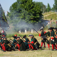 A group of historical reenactors dressed in green and red uniforms, complete with tricorn hats, participate in a battle reenactment reminiscent of the 2010 Dunamunde event. Some soldiers kneel while others stand, aiming long rifles. A large, ornate flag is held by one reenactor as spectators watch from a grassy hill.