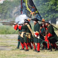 A group of historical reenactors in 18th-century military uniforms fire muskets during a staged battle at Dunamunde 2010. The soldiers wear dark green coats, red breeches, and black tricorn hats. Smoke emerges from the muskets. Trees and stone structures are visible in the background, along with a vibrant flag on display.