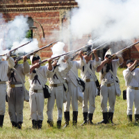 A group of people dressed in historical military uniforms stands in a row on a grassy field, firing old-fashioned muskets. Smoke from the gunfire fills the air. Behind them, there is a brick building with arched windows. The scene, reminiscent of Dunamunde 2010, appears to be a reenactment of a historical battle.