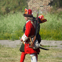 Two individuals dressed in historical military attire stand outdoors. The person in the foreground, reminiscent of Dunamunde reenactors from 2010, wears a red coat, white knee-high socks, and a fur hat with a red emblem. They carry a long rifle on their back. The background shows green grass, a brick pile, and blurred vegetation.