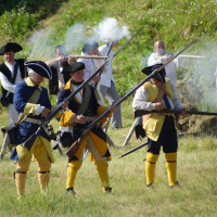 Historical reenactors dressed in 18th-century military uniforms participate in a battle scenario outdoors. Three foreground figures in blue and yellow uniforms are firing muskets, with smoke visible from gunfire, reminiscent of the 2010 Dunamunde event. Background actors in varied attire are engaged, with grassy terrain all around.