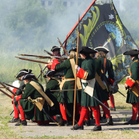 Historical reenactors at Dunamunde 2010 don 18th-century military uniforms, firing muskets and kneeling in formation. They wear dark green coats, red trousers, black tricorn hats, and carry long rifles. Smoke from the muskets fills the air as a complex black and yellow flag with a crest is held by one standing figure.