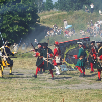 Reenactors in colorful historical military uniforms, with some dressed in yellow and blue and others in red and green, engage in a lively mock battle with muskets and bayonets. Smoke fills the background as a crowd watches from a nearby hill on this sunny 2010 day at Dunamunde.