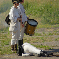 Two soldiers in white uniforms and black hats stand outdoors. One holds a drum, the other a flute. A person in a similar white uniform lies on the ground in front of them, appearing to have fallen, while the soldiers look on. The background features grass and foliage, evoking an eerie scene reminiscent of Dunamunde 2010.