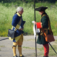 Two people dressed in historical 18th-century European military uniforms are engaged in conversation outdoors. One wears a blue and yellow uniform with a tricorne hat, while the other, donned in green and red, holds a spear in one hand and a white cloth marked "Dunamunde 2010" in the other. They stand on a dirt path with green foliage in the background.