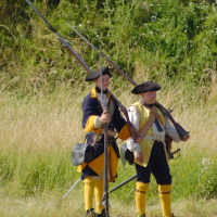 Two reenactors dressed in 18th-century European soldier uniforms stand in a grassy field at Dunamunde 2010. They wear tricorn hats, long blue coats with yellow cuffs, and tall yellow gaiters. Each holds a long musket on their shoulder. A steep grassy hill is visible in the background.