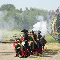 At the 2010 Dunamunde festival, a group of historical reenactors in 18th-century military uniforms fire muskets, creating plumes of smoke. They wear green coats, black tricorn hats, and red stockings. One figure holds a large, decorated flag with a coat of arms. The scene unfolds outdoors amidst greenery and trees.
