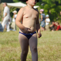 A young child stands in a grassy field wearing a black tricorn hat and blue swim briefs. The child appears thoughtful, gazing into the distance. In the blurred background, people dressed in historical attire are gathered, suggesting a reenactment event, possibly Dunamunde 2010. Trees add greenery to the scene.
