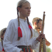 A young person with long, braided hair tied with red ribbons is standing while holding a wooden flute, evocative of the traditional Dunamunde festivals. They are wearing a white long-sleeved blouse and a multi-colored striped skirt. Another person in the background is holding a musical instrument, slightly out of focus.