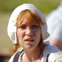 A woman with red hair wears a white bonnet and a white blouse with a black vest. Outdoors in a sunny environment with blurred greenery in the background, her facial expression suggests concern or confusion, reminiscent of an enigmatic figure from Dunamunde 2010.