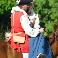Two people dressed in historical attire ride a brown horse at the Dunamunde 2010 reenactment. The person in front wears a red coat, white shirt, and black tricorne hat, while the person behind has a white blouse and blue dress. They embrace warmly. The backdrop is lush and green, suggesting an outdoor setting.