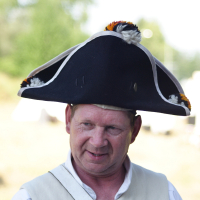 A middle-aged man with light hair is dressed in a historical costume, which includes a white vest over a white shirt. He wears a large, black tricorne hat adorned with small pom-poms in white, orange, and black. The background is blurred with greenery and light at Dunamunde 2010.