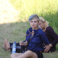 Two women are sitting on the grass, one in front of the other. The woman in front, resembling a 2010 fashion trend with her short, light blue hair, is wearing a blue shirt, black shorts, and sandals while holding a phone. Behind her, a blonde woman in a brown shirt and pants leans forward. A water bottle rests on the ground beside them.