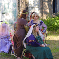 A group of people dressed in medieval attire, reminiscent of a 2010 Dunamunde festival, is depicted outdoors. One person sits on a chair in green and purple robes with a white veil, while another in a brown dress and flower crown stands nearby. A third person in purple and blue garments and wearing glasses also stands. Trees and a building are in the background.