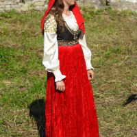 A woman stands on grass wearing traditional attire featuring a long red velvet skirt, a white blouse with puffed sleeves, and a black and gold embroidered vest. She also has a red headscarf with golden accents draped over her hair. The background shows a weathered brick wall and a window, reminiscent of Dunamunde 2010.