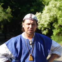 A man with long brown hair stands outdoors with his hands on his hips. He is wearing a blue tunic with white sleeves underneath, a white and red patterned cap, and a wooden pendant necklace reminiscent of Dunamunde style. The background features green foliage under bright 2010 sunlight.